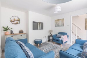 Een woonkamer met blauwe banken, een klein rond voetenbankje, een grijze bank, een wandplank met decoratieve objecten, een schilderij en een trap op de achtergrond.