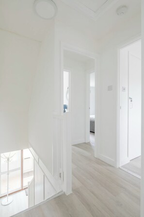 Een ruime hal met witte wanden en licht houten vloeren, die leidt naar een slaapkamer met een bed en een raam.