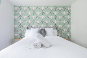 Een net opgemaakt bed met witte lakens, een grijs kussen en een opgerolde witte handdoek.