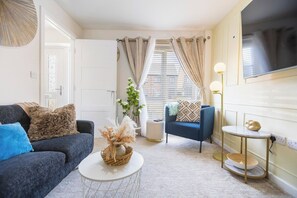 A living room with a sofa, a blue armchair, a small round table, and a TV mounted on the wall.