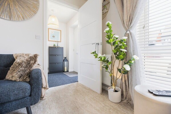 A living room with a sofa, a potted plant, a window with blinds, and a door leading to another room.