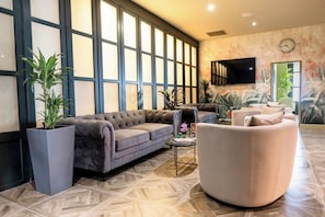 Un hall d’entrée spacieux, doté de grandes fenêtres, d’un canapé gris, d’un fauteuil blanc et de plantes en pot.