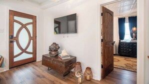 A room with a wooden door, a vintage suitcase, a wall-mounted TV, and a patterned rug. - Walk to Downtown Beaufort | 4BR Coastal Cottage (Beaufort)