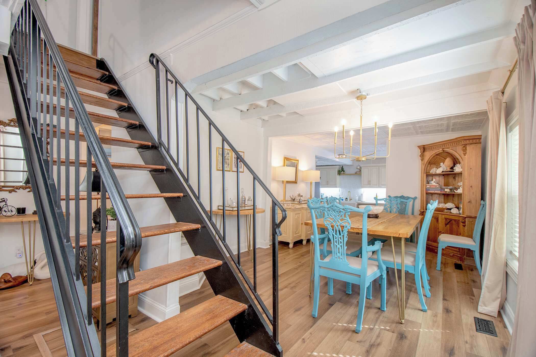Eine Treppe mit Holzboden und schwarzen Geländern, ein Essbereich mit blauen Stühlen und einem Holztisch sowie eine Küche mit weißen Schränken und einer Chandelüster.