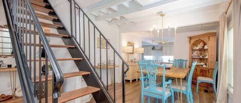Eine Treppe mit Holzboden und schwarzen Geländern, ein Essbereich mit blauen Stühlen und einem Holztisch sowie eine Küche mit weißen Schränken und einer Chandelüster.