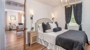 A bedroom with a large bed, bedside lamps, a nightstand, a wooden floor, and a view of a garden through a window. - Walk to Downtown Beaufort | 4BR Coastal Cottage (Beaufort)