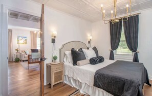 A bedroom with a large bed, bedside lamps, a nightstand, a wooden floor, and a view of a garden through a window. - Charming Coastal Cottage | Walk to Waterfront (Beaufort)