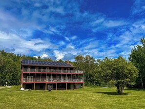 A two-story building with a balcony, solar panels on the roof, and surrounded by a grassy area with trees.