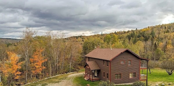 Une cabane en bois dotée d’un porche, entourée d’un feuillage automnal et située au pied d’une colline boisée.