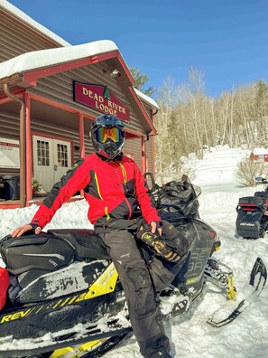 A snowmobile with a rider in a red jacket and helmet, a building with a sign reading 'Dead River Lodge,' and a snowy landscape with trees.