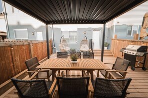 An outdoor patio with a wooden table and chairs, a hanging chair, a grill, and a wooden fence. - Stylish Escape|9 Min to WEM|AC|Gym|Sauna & Patio (Edmonton)