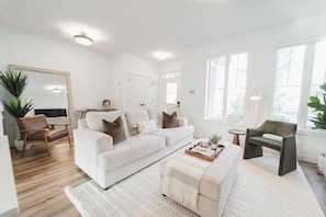A modern living room with a white sofa, a coffee table, and a side table. There is a large mirror, a plant, and a door in the background. - Stylish Escape|9 Min to WEM|AC|Gym|Sauna & Patio (Edmonton)