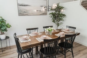A dining room with a wooden table set for six, black chairs, a modern chandelier, a potted plant, and a painting on the wall. - Stylish Escape|9 Min to WEM|AC|Gym|Sauna & Patio (Edmonton)