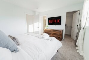 A bedroom with a bed, a TV mounted on the wall, a wooden dresser, and a door leading to another room. - Stylish Escape|9 Min to WEM|AC|Gym|Sauna & Patio (Edmonton)