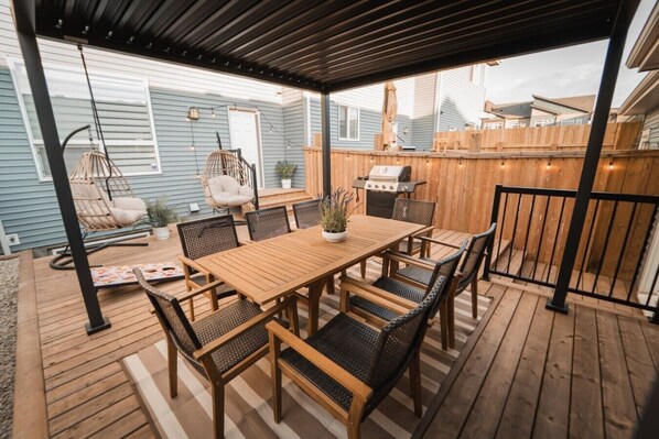 A covered patio with a wooden table and chairs, a hanging chair, a grill, and a wooden fence. - Stylish Escape|9 Min to WEM|AC|Gym|Sauna & Patio (Edmonton)