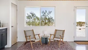 Una habitación con dos sillas de madera, una mesa pequeña, un ventanal con vistas a árboles, una puerta y una zona de cocina.