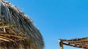 A beach with a pool, a thatched structure, and a building in the background.