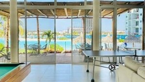 A spacious room with a pool table, a sofa, and large windows offering a view of a pool and resort area.