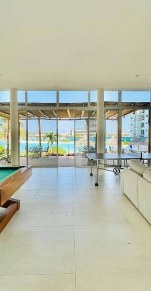 Amplia habitación con mesa de billar, sofá y grandes ventanales que ofrecen vistas a una piscina y al área del resort.