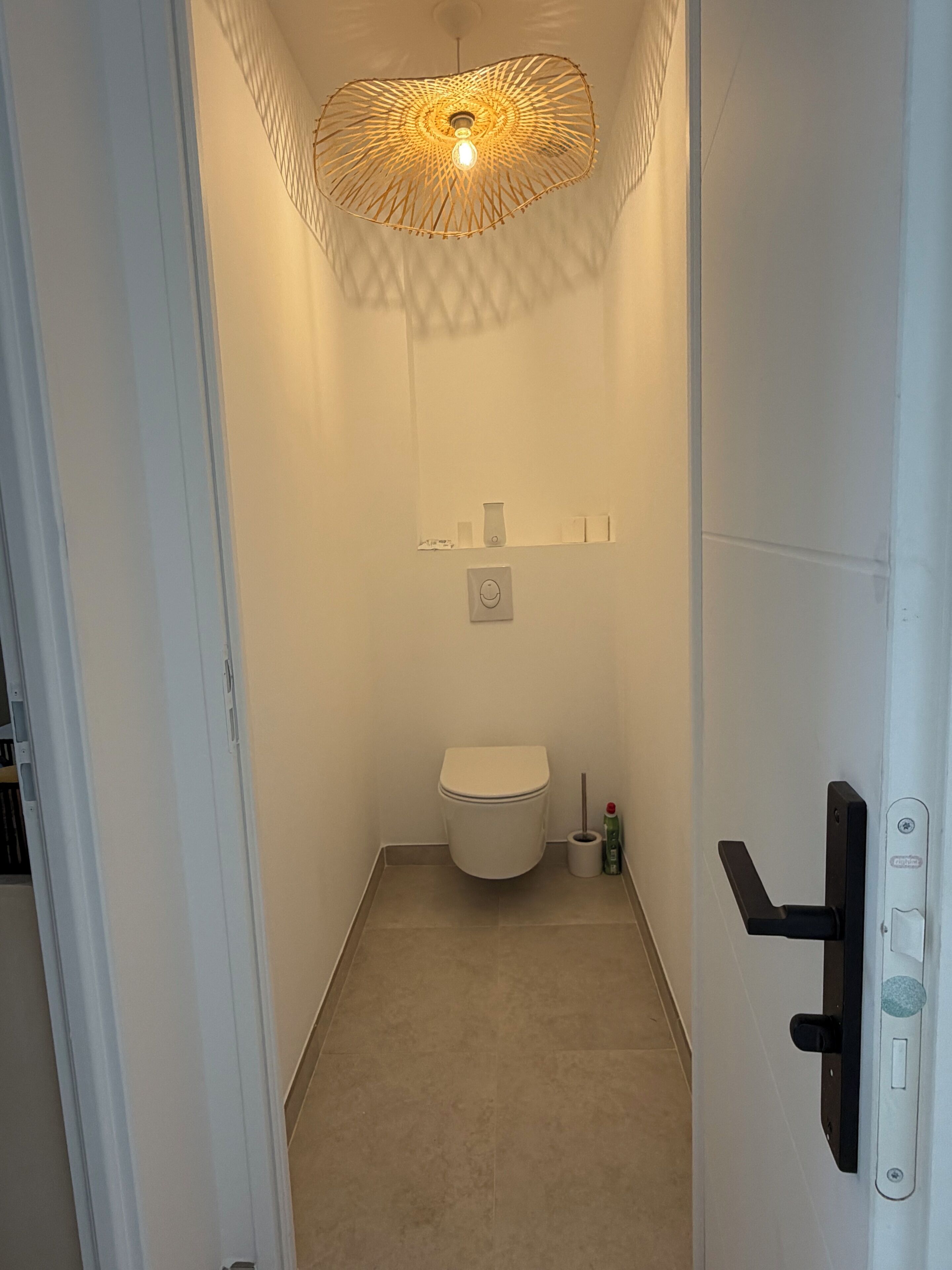 A narrow bathroom with a toilet, a wall-mounted shelf with toiletries, and a unique ceiling light fixture.