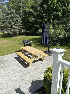 En terrasse med et træbord og grill, omgivet af et hvidt gelænder og grønne planter.