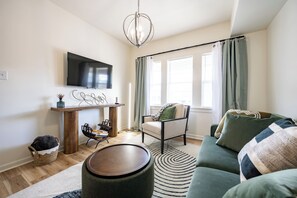 Ein Wohnzimmer mit Flachbildfernseher, Couch, Sessel, Holztisch, Teppich und einem Fenster mit Vorhängen.