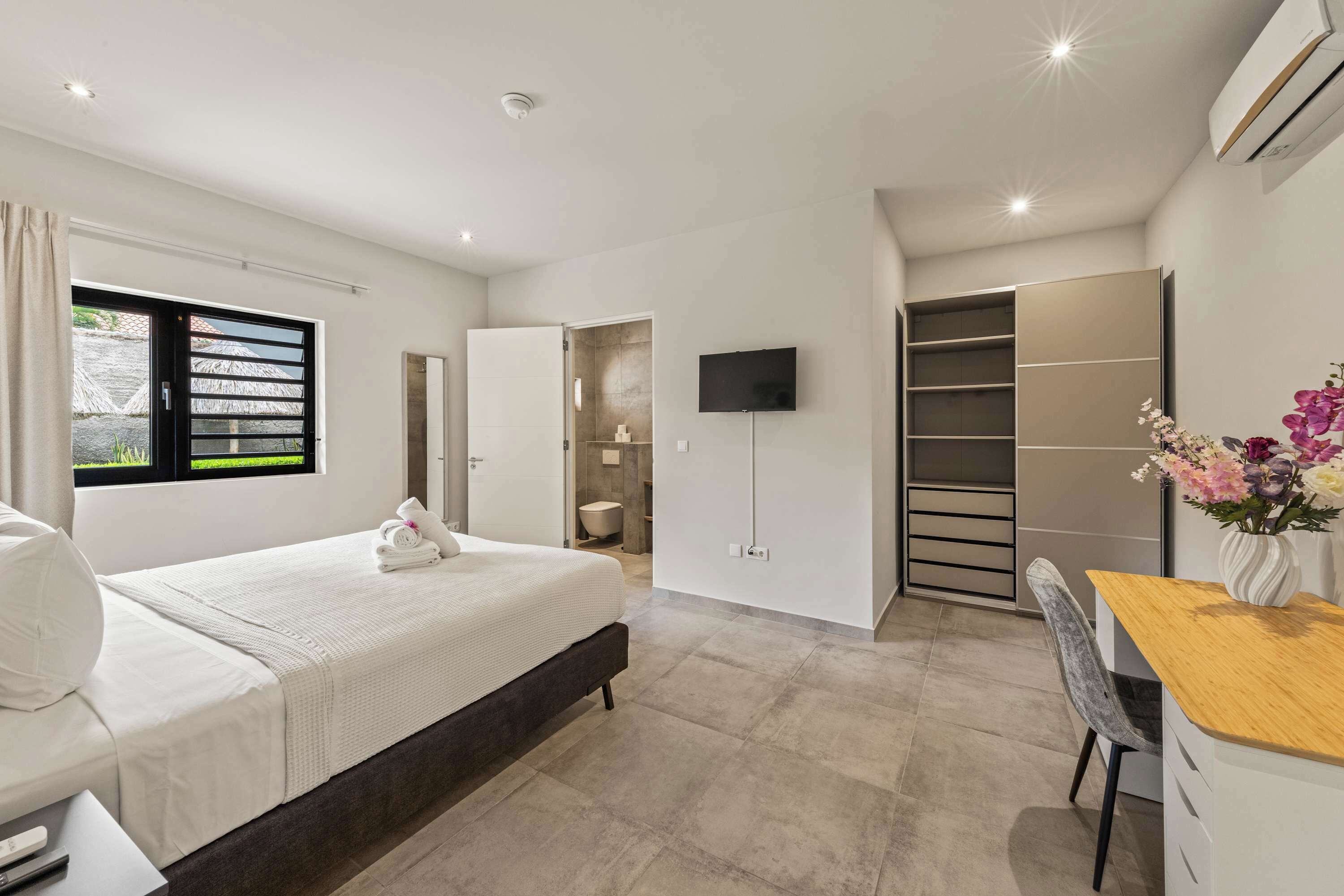 A modern hotel room with a bed, bedside table, chair, TV, wardrobe, and a bathroom visible through an open door.