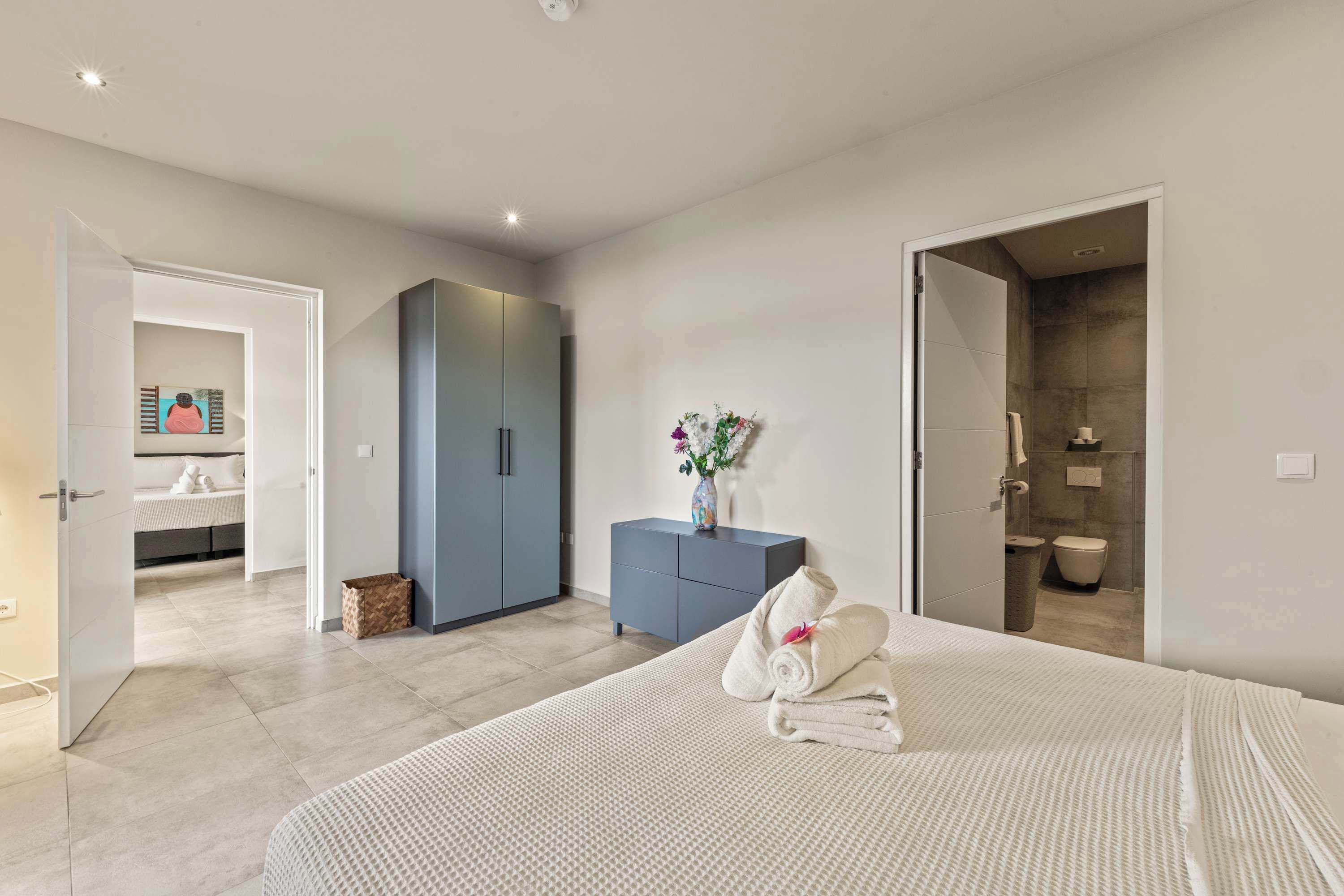 A bedroom with a bed, a dresser, a bathroom visible through an open door, and a hallway leading to another room.