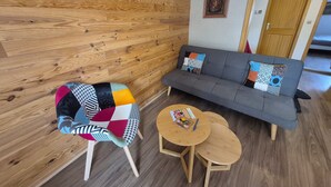 Una habitación con pared de madera, un sofá gris con cojines estampados, una silla de madera con un cojín de retazos y dos pequeñas mesas de madera.