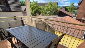 Un balcón con mesa y sillas, barandilla amarilla y vistas a edificios y árboles vecinos.