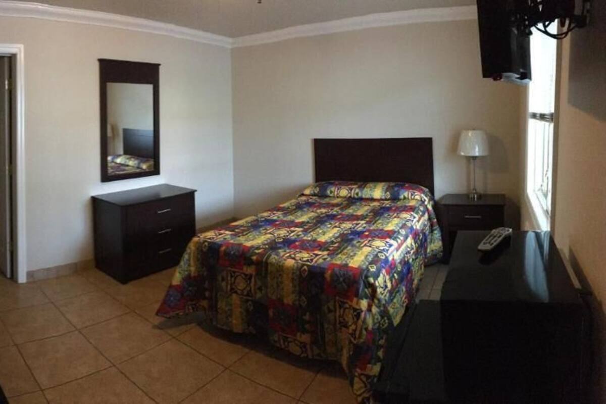 Un dormitorio con cama, cómoda, mesita de noche, espejo, televisor y puerta que da a otra habitación.