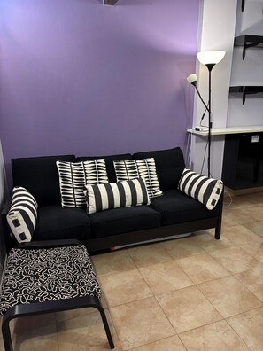 Ein schwarzes Sofa mit gemusterten Kissen, ein passendes Ottoman und eine Stehlampe in einem Zimmer mit violetten Wänden und Fliesenboden.