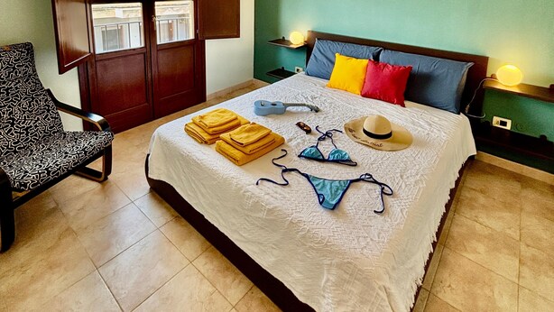 A bedroom with a bed, a chair, a window, and a wall-mounted light fixture. - Calixa - Sheaves - Comfortable apartment for two people, private parking. (Cagliari)