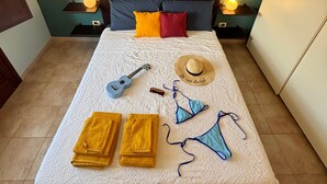 A neatly made bed with a white bedspread, two yellow towels, a blue bikini, a straw hat, a ukulele, and a bottle on the bed. - Calixa - Sheaves - Comfortable apartment for two people, private parking. (Cagliari)