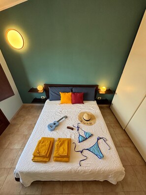 Ein ordentlich gemachtes Bett mit weißer Bettdecke, zwei gelbe Handtücher, einem blauen Bikini, einem Strohhut, einer Ukulele und einer Flasche auf dem Bett.