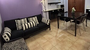 A living room with a black sofa, patterned cushions, a coffee table, a dining table with chairs, a lamp, and a kitchen area with cabinets and a microwave. - Calixa - Sheaves - Comfortable apartment for two people, private parking. (Cagliari)