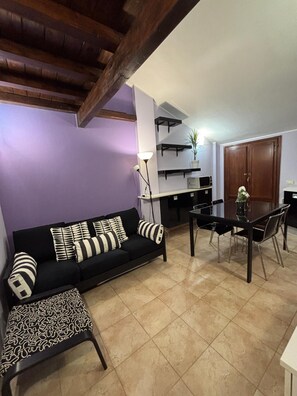 Ein Wohnzimmer mit schwarzem Sofa, gemusterten Kissen, einem Couchtisch, einem Esstisch mit Stühlen, einer Lampe und einer Küchenzeile mit Schränken und einer Mikrowelle.