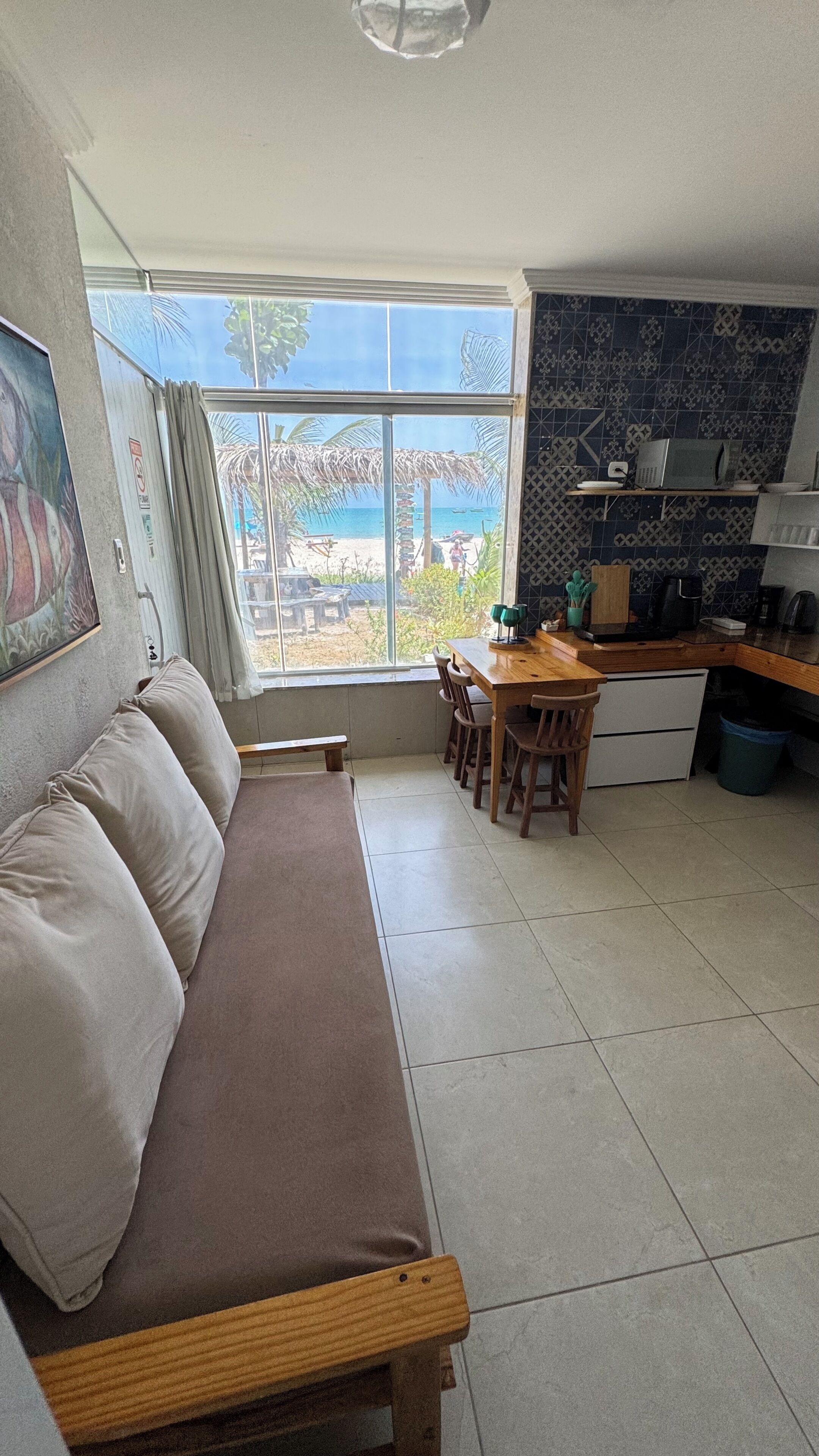Ein Wohnzimmer mit einer Couch, einem Esstisch mit Stühlen, einer Küchenzeile mit Arbeitsfläche und Geräten sowie einem großen Fenster mit Blick auf den Strand.