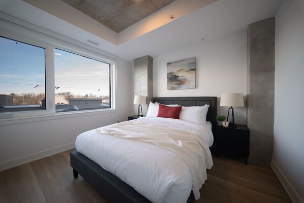 A modern bedroom with a large bed, a nightstand, a lamp, and a window with a view.