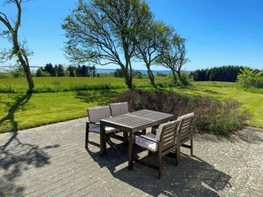 Eine Terrasse mit Tisch und Stühlen, umgeben von Grünpflanzen und Blick auf das Meer.