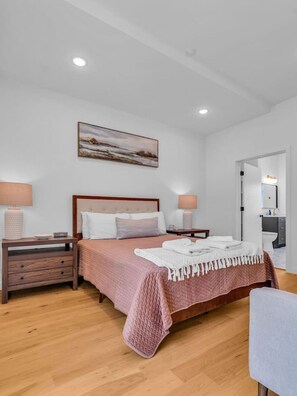 Une chambre à coucher avec un lit, des tables de chevet, une commode, un tableau et une salle de bain visible à travers une porte ouverte.