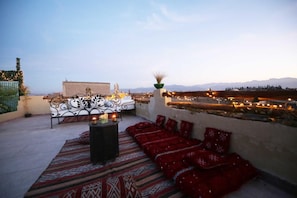 A rooftop terrace with a patterned rug, red cushions, a metal table with candles, and a view of distant mountains. - Riad Étoile du Sud (Sidi Abdallah Ghiat)