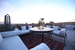 Een terras op het dak met een ronde tafel, wit gestoffeerde zitplaatsen en een kamerkleed met een patroon.