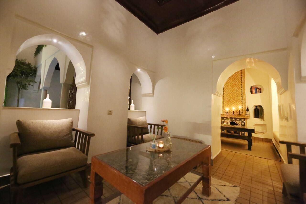 Una habitación con una mesa de vidrio, una silla acolchada y un banco. Se observan puertas abovedadas y un suelo de baldosas.