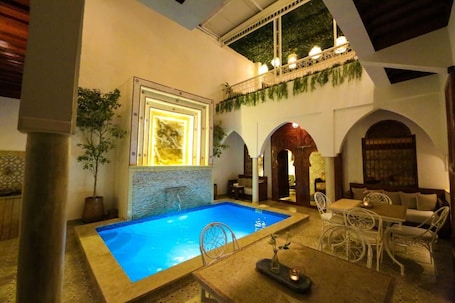 An indoor swimming pool with a tiled floor, surrounded by white wicker furniture and a dining area with a table and chairs.. Riad Étoile du Sud