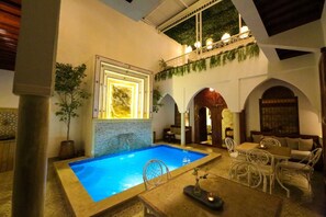 An indoor swimming pool with a tiled floor, surrounded by white wicker furniture and a dining area with a table and chairs. - Riad Étoile du Sud (Sidi Abdallah Ghiat)