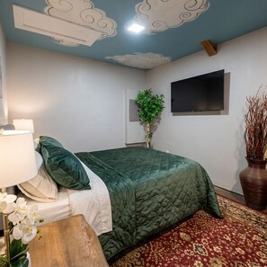 Una camera da letto con un letto, un comodino, una lampada, una pianta, una TV e un soffitto decorato.