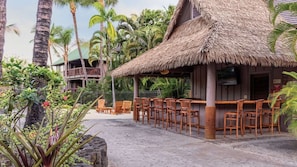 A resort with a thatched-roof bar area, wooden chairs, and a view of palm trees and a building with balconies.