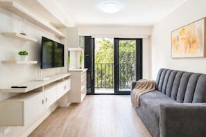 Ein modernes Wohnzimmer mit Flachbildfernseher, Einbauschränken, einem grauen Sofa und einem Balkon mit Blick auf Grünflächen.