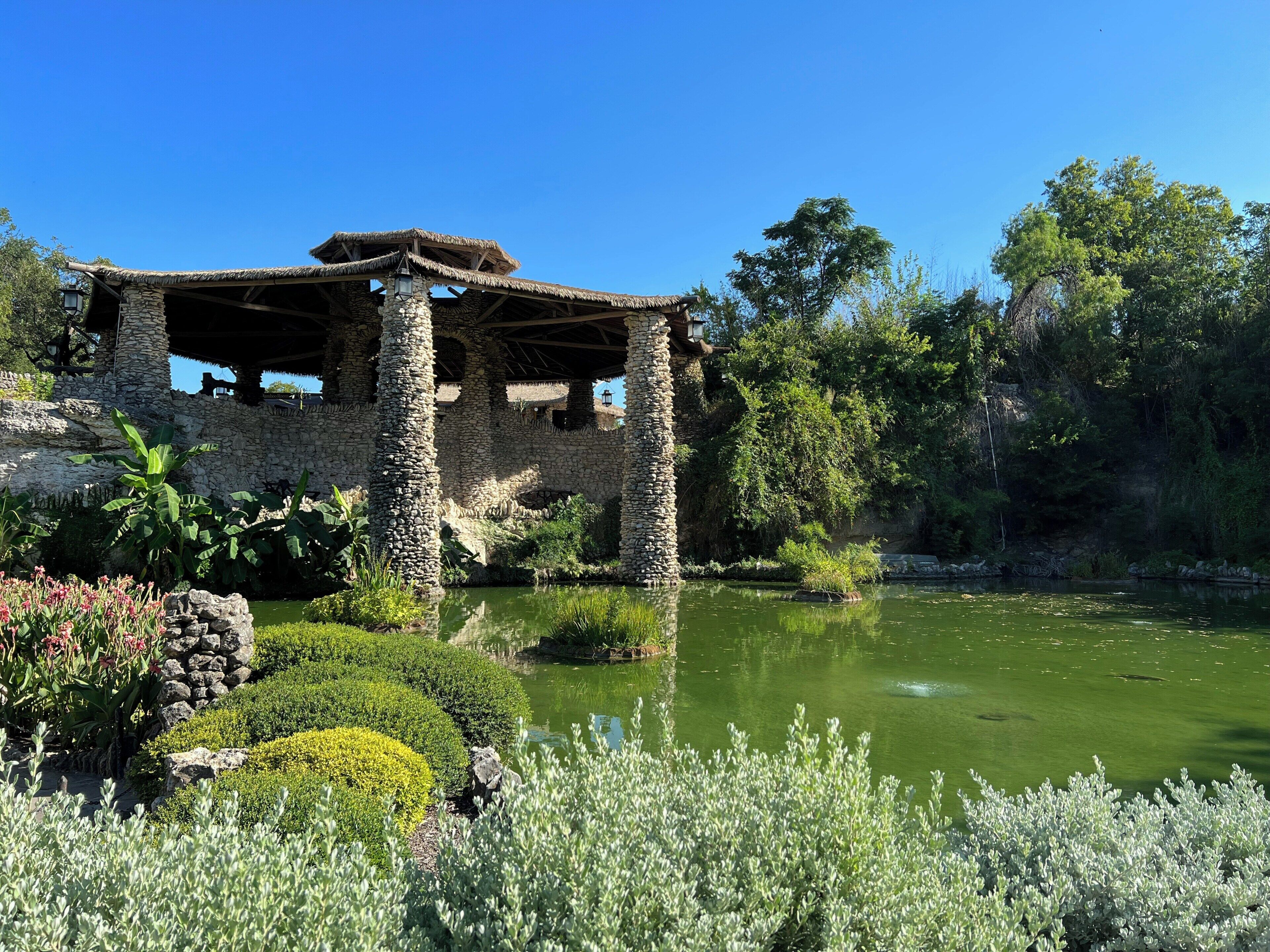 A stone gazebo with a thatched roof, surrounded by lush greenery and a pond.